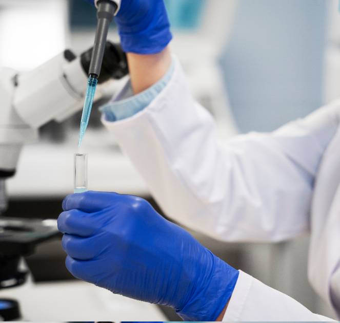 Scientist in white lab coat and blue gloves carefully using a pipette to transfer liquid into a test tube beside a microscope in a modern clinical laboratory