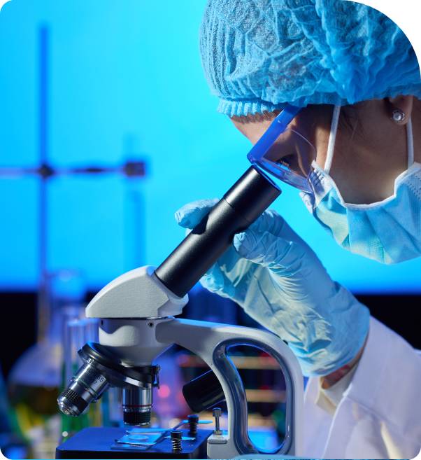 Scientist wearing blue lab coat, gloves, surgical cap, and face mask looking through a microscope in a modern clinical laboratory with blue lighting